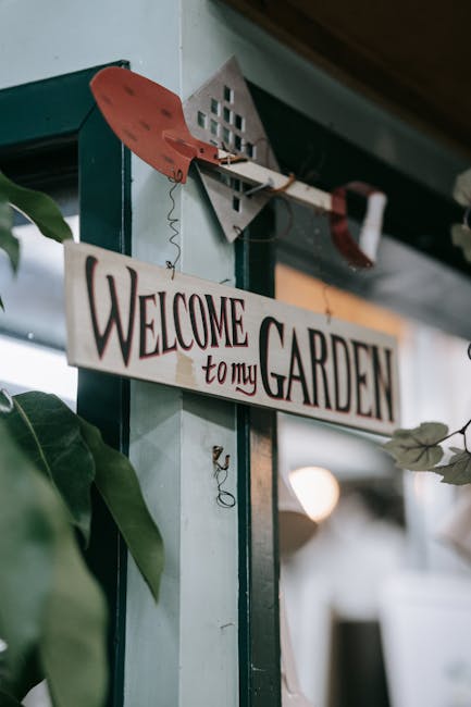 A close-up view of a wooden sign hanging inside a garden center or outdoor space, with the words 'Welcome to my garden' painted in decorative black and red lettering. The sign is attached to a light b