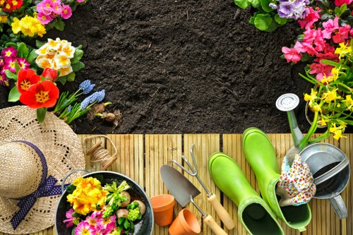 The image shows a neatly organized garden edging scene with a wooden patio area bordered by fresh, dark soil ready for planting. To the left, there is a variety of colourful flowering plants, including pink, purple, yellow, and red blooms, creating a vibrant contrast against the dark earth. The foreground features gardening tools and accessories arranged on the wooden decking, including a pair of bright green rubber boots, a metal watering can with a floral design, small terracotta pots, and a black plastic bucket filled with potted flowering plants. A wide-brimmed straw hat with a purple ribbon rests on the edge of the decking, providing a touch of outdoor gardening readiness. In the background, the garden extends into more flower beds and possibly hedges or bushes, indicating a well-maintained outdoor space suitable for gardening and landscaping services. The scene appears to be set on a bright, clear day with natural daylight illuminating the vibrant colours and textures, emphasizing a productive and inviting garden environment that aligns with professional gardening care offered by Gardener Belmont in the Belmont area near London.