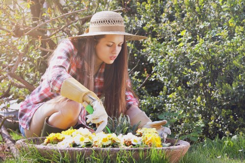 A young woman with long brown hair wearing a wide-brimmed straw hat, red and black plaid shirt, and gardening gloves is kneeling in a lush garden. She is tending to a flower bed filled with yellow and white flowering plants, using her hands to carefully arrange or extract plants. Behind her, there are dense green bushes and trees, with sunlight filtering through the foliage, creating a natural and vibrant outdoor scene. The garden features a grass lawn and a circular border around the flower bed, reflecting a well-maintained landscaped yard. This scene emphasizes outdoor gardening activities typical of professional garden care services provided by Gardener Belmont in the Belmont area, supporting local landscaping and lawn maintenance. The environment suggests a peaceful, sunny day with warm weather, ideal for garden work, and highlights the beauty of a well-kept garden space for homeowners in the region.